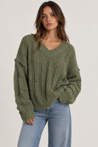 Model wearing an oversized textured cable-knit V-neck sweater in olive, featuring an open-knit design, dropped shoulders, long sleeves, and ribbed trim, styled with light-wash denim.
