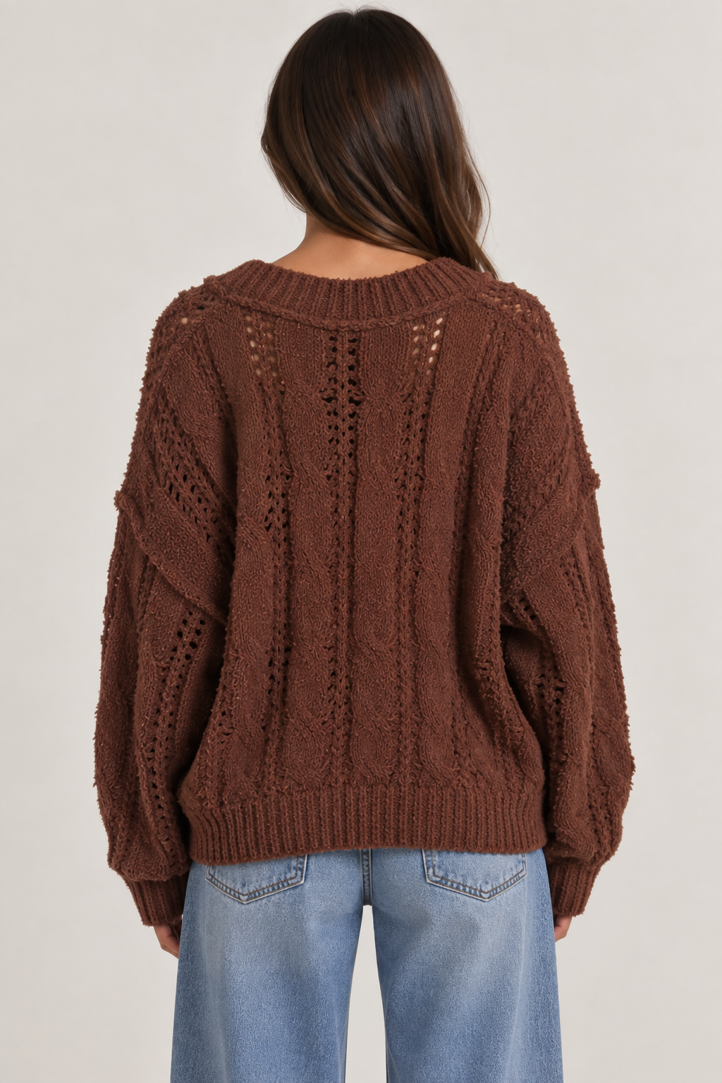 Model wearing an oversized textured cable-knit V-neck sweater in brown, featuring an open-knit design, dropped shoulders, long sleeves, and ribbed trim, styled with light-wash denim.
