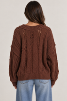 Model wearing an oversized textured cable-knit V-neck sweater in brown, featuring an open-knit design, dropped shoulders, long sleeves, and ribbed trim, styled with light-wash denim.