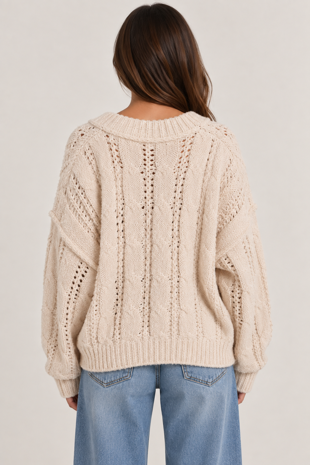 Model wearing an oversized textured cable-knit V-neck sweater in cream, featuring an open-knit design, dropped shoulders, long sleeves, and ribbed trim, styled with light-wash denim.