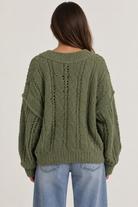 Model wearing an oversized textured cable-knit V-neck sweater in olive, featuring an open-knit design, dropped shoulders, long sleeves, and ribbed trim, styled with light-wash denim.