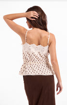 Cream camisole top with a subtle brown polka dot print, scalloped floral lace trim, adjustable spaghetti straps, and a gathered tie waist. A feminine, romantic style with a softly flared hem and delicate lace details.
