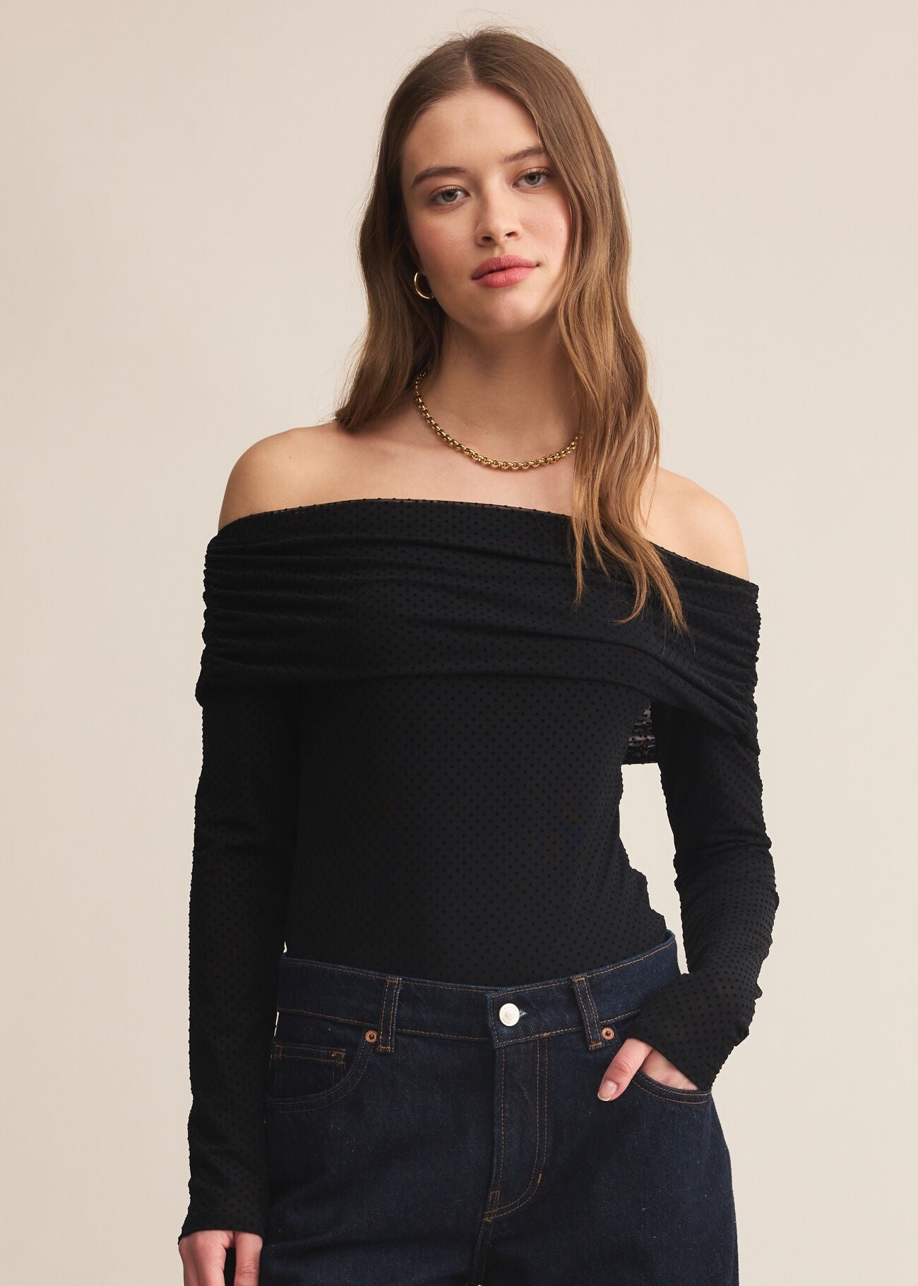Woman wearing a black off-shoulder top and dark jeans on a beige background