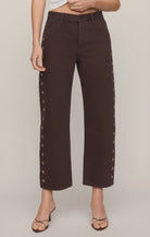 Rich brown mid-rise denim jeans featuring a relaxed bowed-leg silhouette, ankle-length hem, gold grommet details, and subtle side openings.
